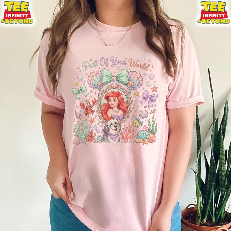 May include: A light pink t-shirt featuring a colorful graphic of Ariel from The Little Mermaid. The design includes the text "Part Of Your World", a portrait of Ariel, a dog, and various sea-themed elements like seashells and coral.