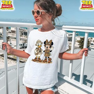 May include: White t-shirt featuring a cartoon illustration of Donald Duck and Minnie Mouse in safari outfits. Donald Duck is wearing a khaki vest and hat, while Minnie Mouse sports a leopard print outfit. The shirt has the text "TEE INFINITY & BEYOND" at the top.