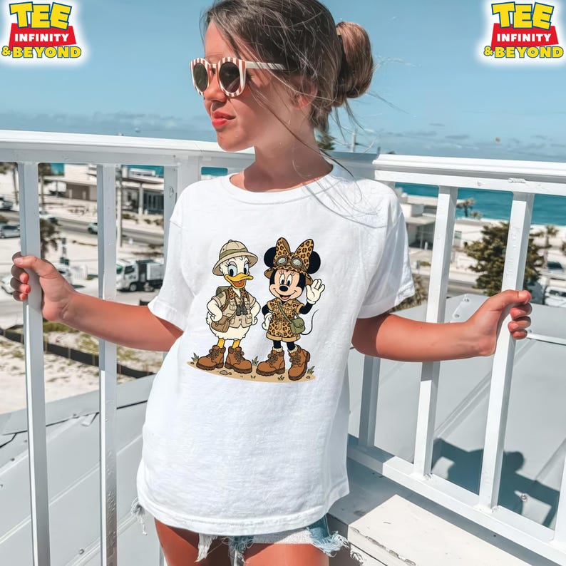 May include: White t-shirt featuring a cartoon illustration of Donald Duck and Minnie Mouse in safari outfits. Donald Duck is wearing a khaki vest and hat, while Minnie Mouse sports a leopard print outfit. The shirt has the text "TEE INFINITY & BEYOND" at the top.