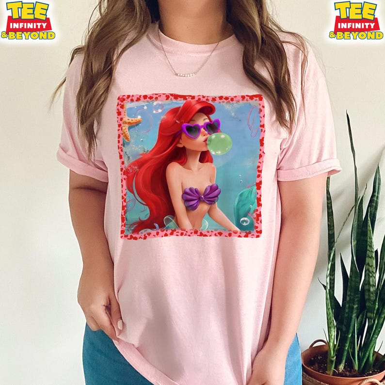 May include: A light pink t-shirt featuring a graphic of a cartoon mermaid with red hair, purple sunglasses, and a bubble. The mermaid is wearing a purple seashell top. The shirt has the text "TEE INFINITY & BEYOND" on the top left and right.