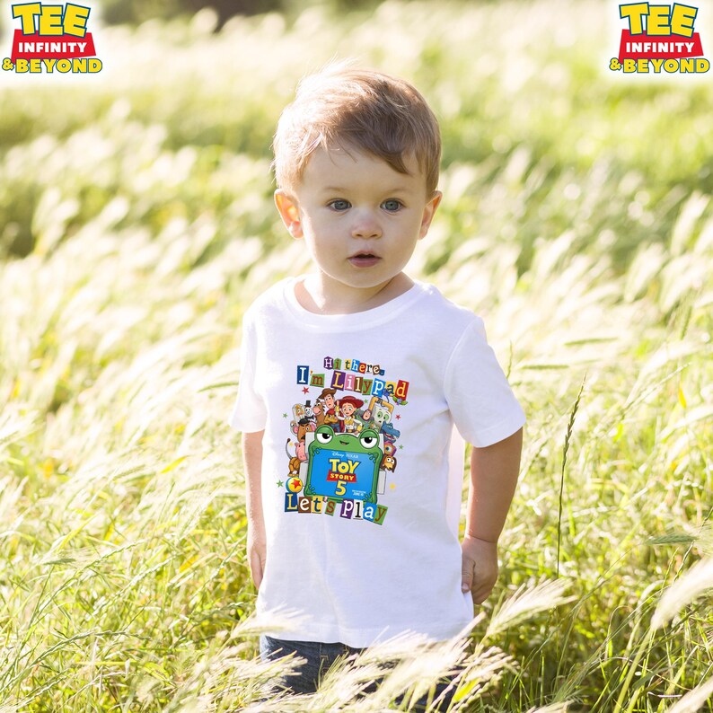 May include: A white short-sleeved t-shirt featuring a colorful graphic print of Toy Story characters, a green frog illustration, and the text Hi there, I'm Lilypad, Toy Story 5, Let's Play. The shirt has a crew neckline and a standard fit design.