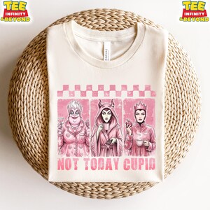 May include: Cream-colored t-shirt featuring three cartoon villains in pink frames. The shirt has the text "NOT TODAY CUPID" in pink. The villains are Ursula, Maleficent, and the Evil Queen. The shirt is on a woven surface.