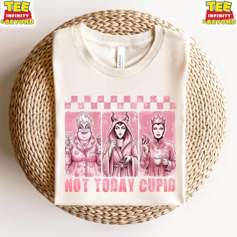 May include: Cream-colored t-shirt featuring three cartoon villains in pink frames. The shirt has the text "NOT TODAY CUPID" in pink. The villains are Ursula, Maleficent, and the Evil Queen. The shirt is on a woven surface.
