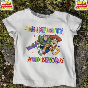 May include: White t-shirt with a colourful graphic of Buzz Lightyear and Woody from Toy Story. The text "TO INFINITY AND BEYOND" is written above and below the characters in rainbow colours. The shirt has short sleeves.