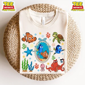 May include: A light beige t-shirt with a colorful cartoon design featuring Dory, Nemo, and other characters from the movie Finding Nemo. The design includes a blue and yellow fish, a green turtle, a red octopus, and a pink jellyfish. The shirt has the words "Tee Infinity & Beyond" in the top left and top right corners.