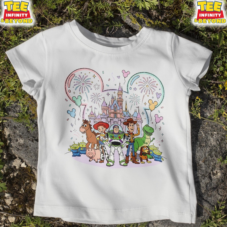 May include: White t-shirt featuring a colorful graphic of characters from the Toy Story movie, including Woody, Buzz Lightyear, and others, in front of a castle and fireworks display. The shirt also has the text "TEE INFINITY & BEYOND".