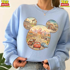 May include: Light blue sweatshirt featuring a Mickey Mouse head design with a Cars theme. The design includes a race track, desert landscape, and Route 66 elements. The sweatshirt has the text "TEE INFINITY & BEYOND" on the top left and right.