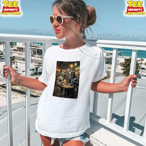 K&ouml;nnte beinhalten: Wei&szlig;es T-Shirt mit einem Star Wars-Motiv von Yoda und Darth Vader in einem Pub. Das T-Shirt hat oben den Text "TEE INFINITY & BEYOND". Das Bild ist auf einer Person mit Sonnenbrille, die sich drau&szlig;en aufh&auml;lt.