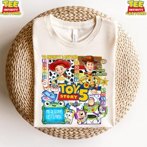 May include: A cream-colored t-shirt featuring a colorful Toy Story 5 graphic. The design includes characters Jessie, Woody, and Buzz Lightyear, along with text that reads To Infinity & Beyond, It's Called A Poncho, and I'm Always Listening.