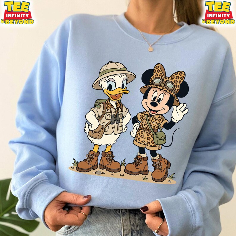 May include: Light blue sweatshirt featuring cartoon characters Donald Duck and Minnie Mouse dressed in safari outfits. Donald Duck wears a khaki safari hat, vest, and boots. Minnie Mouse is in a leopard print outfit with a matching bow and hat. The shirt has the text "TEE INFINITY & BEYOND".
