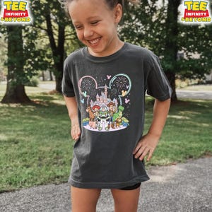 May include: Dark gray t-shirt featuring a colorful graphic of Disney's Toy Story characters in front of a castle and fireworks display. The shirt has the text "TEE INFINITY & BEYOND" in yellow. The shirt is worn by a young person.