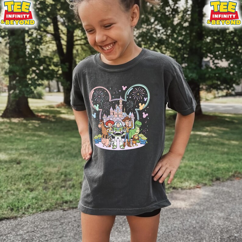 May include: Dark gray t-shirt featuring a colorful graphic of Disney's Toy Story characters in front of a castle and fireworks display. The shirt has the text "TEE INFINITY & BEYOND" in yellow. The shirt is worn by a young person.