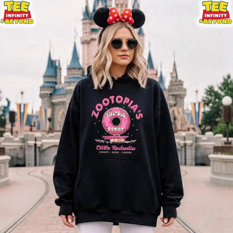 Puede incluir: Sudadera negra con un gr&aacute;fico de donut rosa y el texto "ZOOTOPIA'S THE BIG DONUT" y "Cittle Rodentia". La sudadera tiene un bolsillo delantero y una capucha con cord&oacute;n. Un castillo se ve en el fondo.