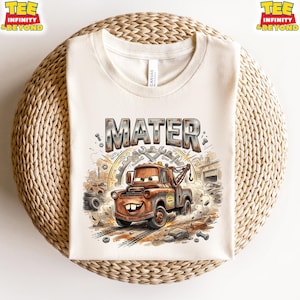 May include: Cream-colored t-shirt featuring a cartoon image of Mater, the tow truck from the movie Cars. The word "MATER" is printed above the image in a distressed, metallic font. The design includes tools, gears, and a background of a garage setting.
