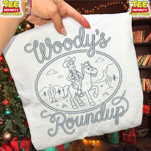 May include: A white t-shirt featuring a dark blue line art graphic of a cowboy riding a horse inside a rope lasso frame. The text "Woody's Roundup" is written in a rope-style font. The design includes desert elements like cacti and clouds.