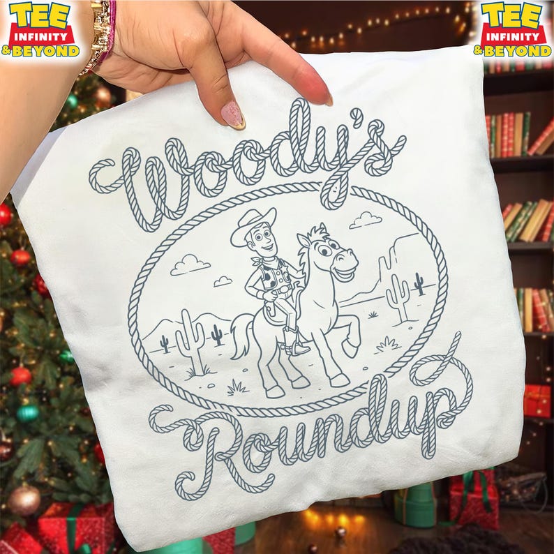 May include: A white t-shirt featuring a dark blue line art graphic of a cowboy riding a horse inside a rope lasso frame. The text "Woody's Roundup" is written in a rope-style font. The design includes desert elements like cacti and clouds.