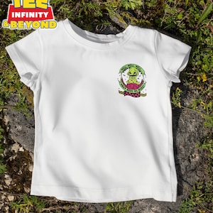 May include: White t-shirt with a graphic featuring a green creature with the text "Well, Well, Well, What Have We Here?". The design includes a pink and black checkered pattern. The shirt is displayed on a grassy surface.