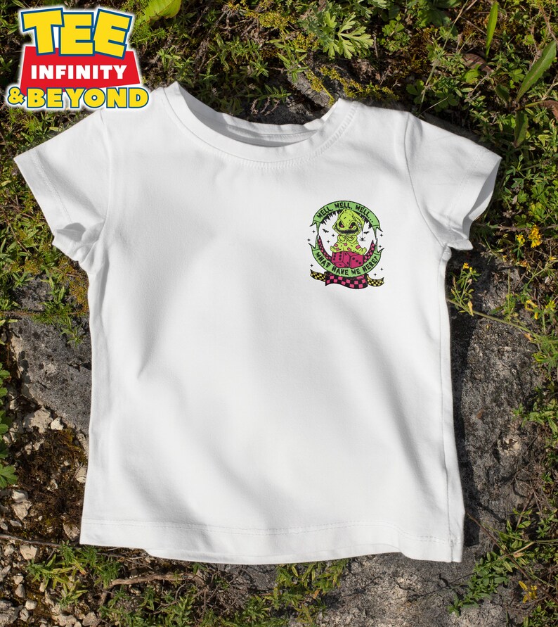 May include: White t-shirt with a graphic featuring a green creature with the text "Well, Well, Well, What Have We Here?". The design includes a pink and black checkered pattern. The shirt is displayed on a grassy surface.