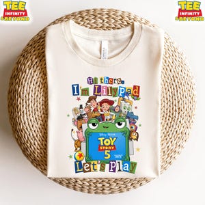 May include: A cream-colored t-shirt featuring a colorful graphic of Toy Story characters surrounding a green frog-shaped frame. The text reads Hi there, I'm Lilypad, Toy Story 5, and Let's Play. The design includes various animated figures.
