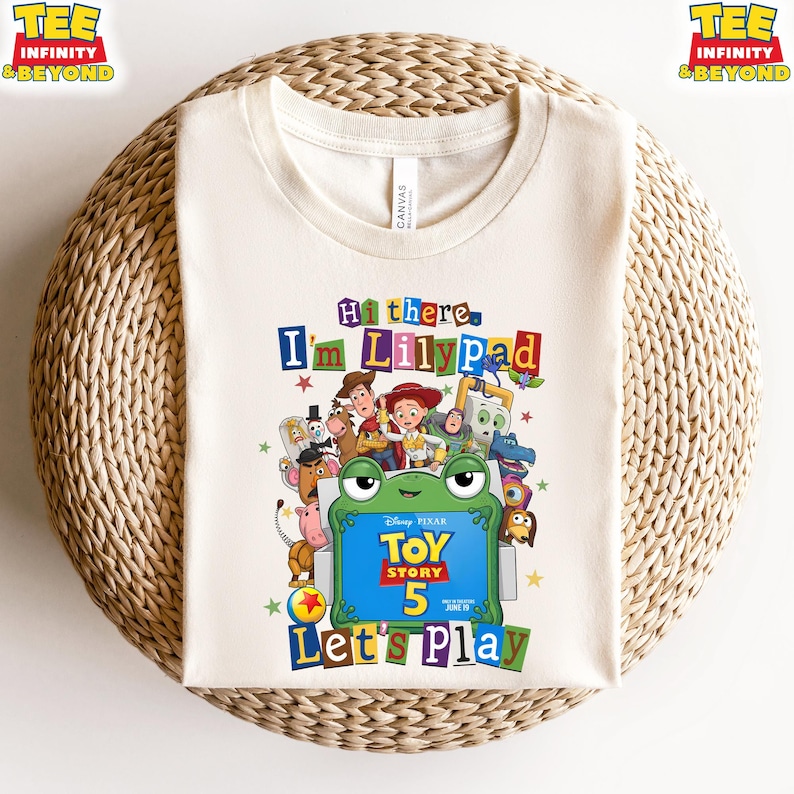 May include: A cream-colored t-shirt featuring a colorful graphic of Toy Story characters surrounding a green frog-shaped frame. The text reads Hi there, I'm Lilypad, Toy Story 5, and Let's Play. The design includes various animated figures.