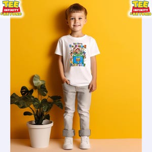 May include: A white short-sleeved t-shirt featuring a colorful graphic print of various animated characters, a green frog, and the text "I'm Toypad Let's Play" along with "Toy Story 5". The shirt is paired with light gray denim pants and white sneakers.
