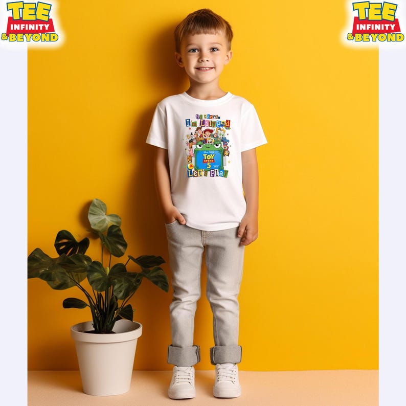 May include: A white short-sleeved t-shirt featuring a colorful graphic print of various animated characters, a green frog, and the text "I'm Toypad Let's Play" along with "Toy Story 5". The shirt is paired with light gray denim pants and white sneakers.