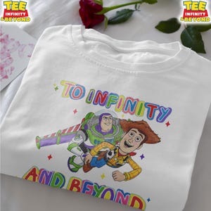 May include: White t-shirt with a colourful graphic of Buzz Lightyear and Woody from Toy Story, and the text "TO INFINITY AND BEYOND" in rainbow colours. The shirt has a crew neck and short sleeves.