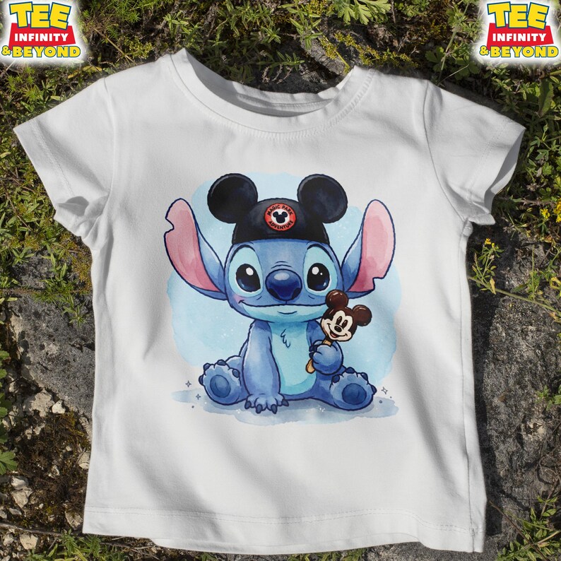 K&ouml;nnte beinhalten: Wei&szlig;es T-Shirt mit einer blauen Cartoonfigur, die einen schwarzen Hut mit Mickey Mouse-Ohren tr&auml;gt und ein Mickey Mouse-Eis h&auml;lt. Das T-Shirt hat den Text "TEE INFINITY & BEYOND" oben links und rechts.