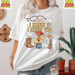 May include: A white t-shirt featuring a graphic print of a cowgirl character with a blonde braid, wearing a red hat, yellow shirt, and blue jeans. The text reads "I KNEW IT I KNEW YOU", "YOU'VE GOT A FRIEND IN ME", "SHERIFF", and "It's a Jessie Story Baby Just Say Yes".