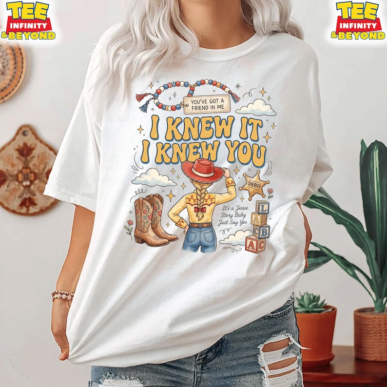 May include: A white t-shirt featuring a graphic print of a cowgirl character with a blonde braid, wearing a red hat, yellow shirt, and blue jeans. The text reads "I KNEW IT I KNEW YOU", "YOU'VE GOT A FRIEND IN ME", "SHERIFF", and "It's a Jessie Story Baby Just Say Yes".