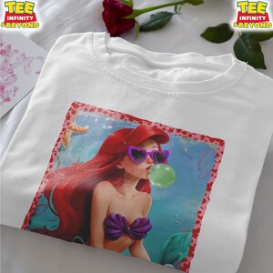 May include: White t-shirt featuring a colorful graphic of a mermaid with red hair, wearing heart-shaped sunglasses and blowing a bubble. The mermaid is in a blue underwater scene. The shirt has the text "TEE INFINITY & BEYOND" on the top.