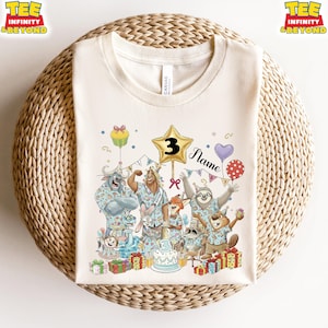 Puede incluir: Camiseta color crema con un dise&ntilde;o de cumplea&ntilde;os colorido. El gr&aacute;fico incluye personajes de animales de dibujos animados, una estrella dorada con el n&uacute;mero 3, globos, una tarta y la palabra "Name". La camiseta tiene el texto "TEE INFINITY & BEYOND" en las esquinas superiores.