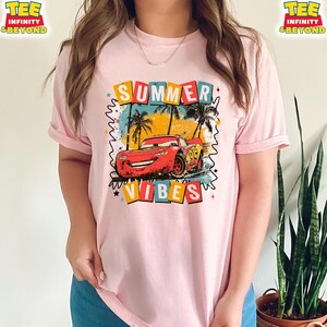 May include: Pink t-shirt featuring a graphic of a red race car with the text "SUMMER VIBES". The design includes palm trees and colorful text blocks. The shirt has short sleeves.
