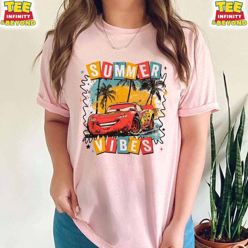 May include: Pink t-shirt featuring a graphic of a red race car with the text "SUMMER VIBES". The design includes palm trees and colorful text blocks. The shirt has short sleeves.