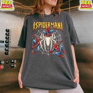 May include: Dark gray t-shirt featuring a graphic of Spider-Man in action poses with a spider web background. The text "SPIDER-MAN" is displayed in yellow with a lightning bolt design. The shirt has the text "TEE INFINITY & BEYOND" on the top right.