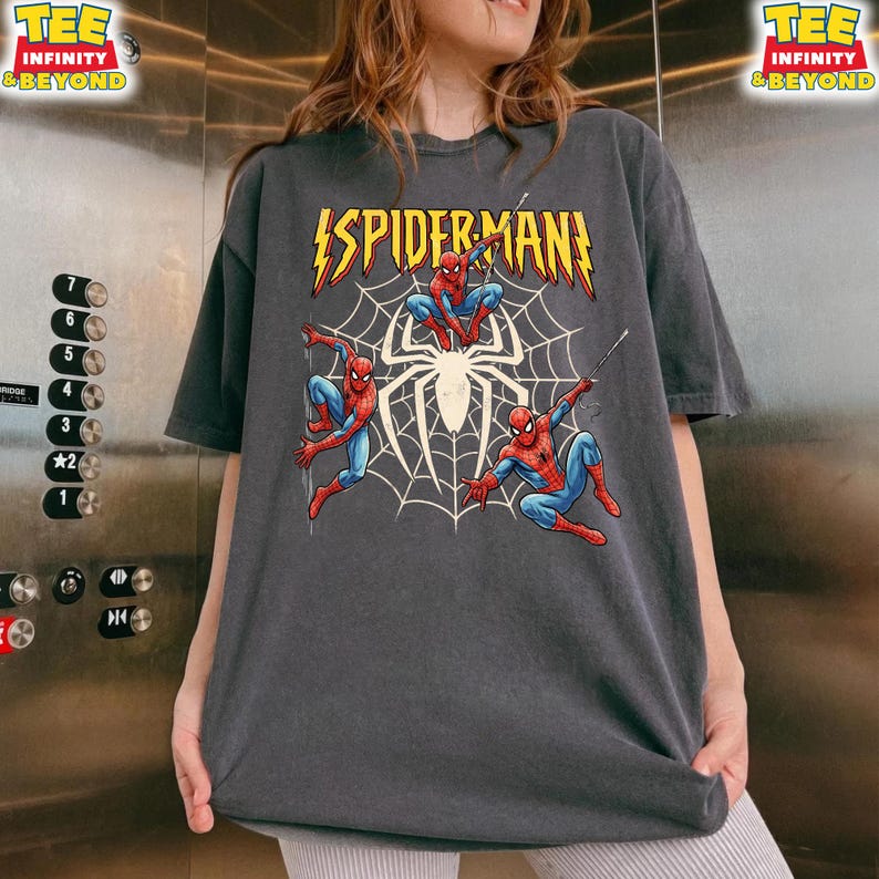 May include: Dark gray t-shirt featuring a graphic of Spider-Man in action poses with a spider web background. The text "SPIDER-MAN" is displayed in yellow with a lightning bolt design. The shirt has the text "TEE INFINITY & BEYOND" on the top right.