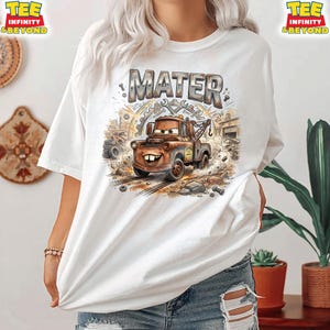 May include: White t-shirt featuring a cartoon illustration of Mater from the Cars movie. The design includes the name "MATER" in a distressed font, along with tools and a tow truck. The shirt has short sleeves and a crew neck.