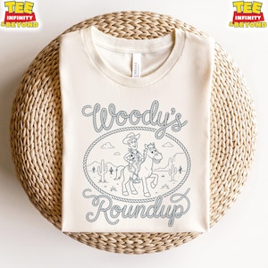 May include: A cream-colored crew neck t-shirt featuring a dark blue line art graphic of a cowboy riding a horse inside a lasso rope frame. The text Woody's Roundup is written in a rope-style font above and below the central illustration.