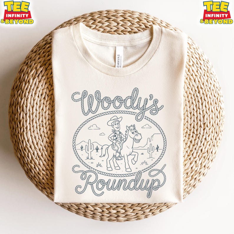 May include: A cream-colored crew neck t-shirt featuring a dark blue line art graphic of a cowboy riding a horse inside a lasso rope frame. The text Woody's Roundup is written in a rope-style font above and below the central illustration.