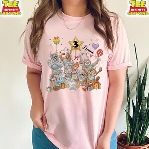 Puede incluir: Camiseta rosa claro con un dise&ntilde;o colorido de animales de dibujos animados celebrando un cumplea&ntilde;os. El dise&ntilde;o incluye un globo en forma de estrella con el n&uacute;mero 3, una tarta y la palabra "Name". La camiseta tiene mangas cortas y un corte holgado.