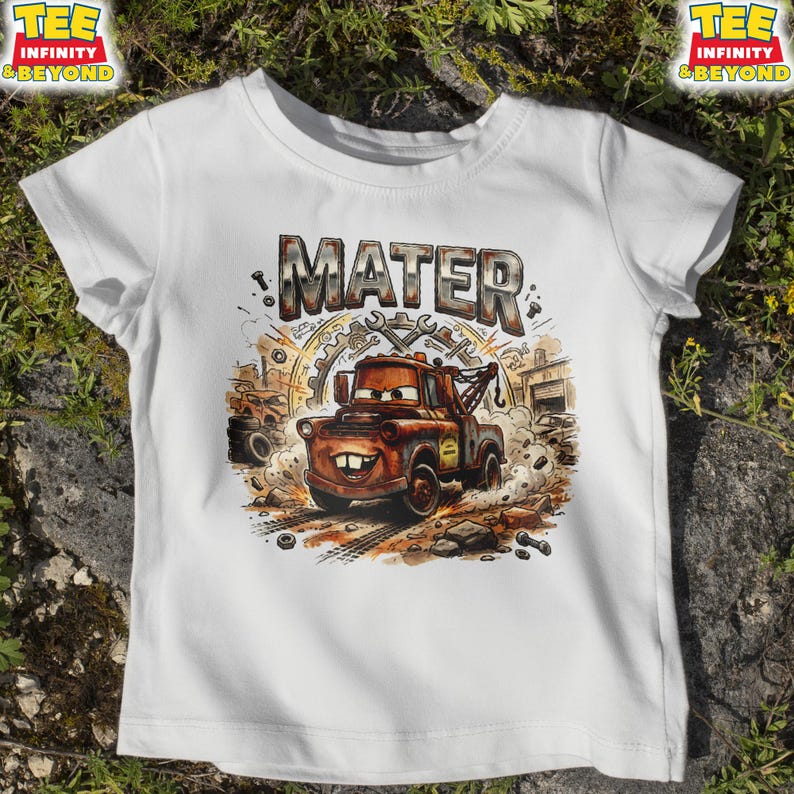 May include: White t-shirt featuring a cartoon illustration of Mater from the Cars movie. The word "MATER" is printed above the character in a distressed, metallic font. The design includes a detailed background with tools and a garage setting.