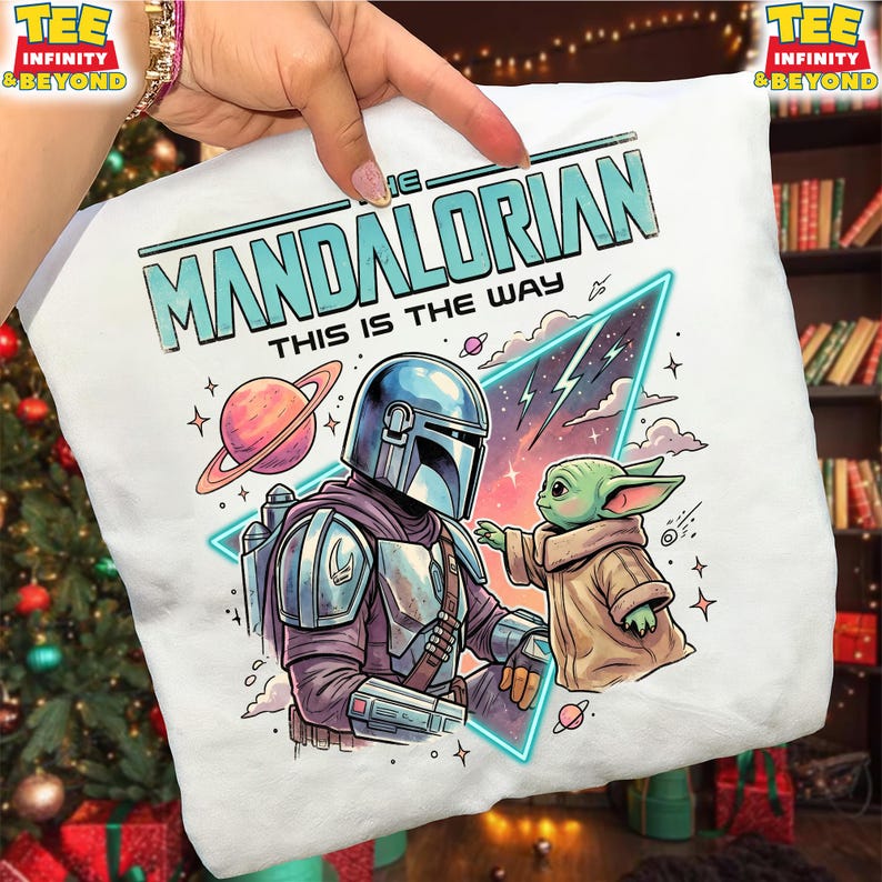 Może przedstawiać: Biały t-shirt z nadrukiem graficznym przedstawiającym Mandalorianina i Grogu. Tekst głosi THE MANDALORIAN THIS IS THE WAY. Wz&oacute;r zawiera planetę z pierścieniami, tr&oacute;jkątną neonową ramkę oraz gwiazdy na jasnym tle.