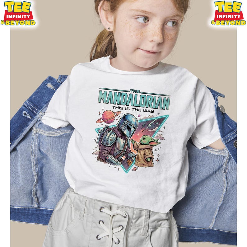 Może przedstawiać: Biały t-shirt z kr&oacute;tkim rękawem z grafiką przedstawiającą Mandalorianina i Grogu. Napis głosi THE MANDALORIAN THIS IS THE WAY. Wz&oacute;r zawiera planetę, gwiazdy i tr&oacute;jkątną ramkę. Koszulka jest założona pod niebieską kurtkę dżinsową.