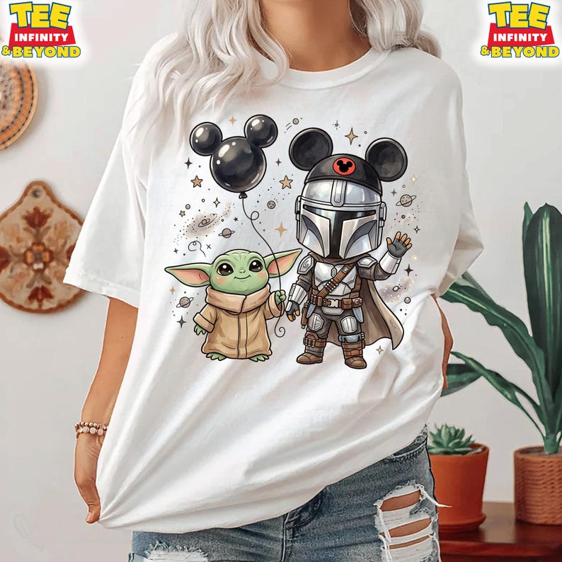 May include: A white t-shirt featuring a graphic of a small green creature in a tan robe and a silver-armored character wearing black mouse ears. The design includes a black balloon, small stars, and planets. The text Tee Infinity & Beyond is visible.