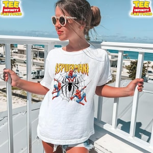 May include: White t-shirt featuring a Spider-Man graphic. The design includes the word "SPIDERMAN" in yellow and red, along with illustrations of Spider-Man and a black spider web. The shirt is worn by a person outdoors.