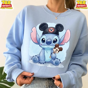 K&ouml;nnte beinhalten: Hellblaues Sweatshirt mit einer Cartoon-Illustration von Stitch, der einen Mickey Mouse-Hut tr&auml;gt und einen Mickey Mouse-Eis am Stiel h&auml;lt. Der Hut tr&auml;gt den Text "Magic Park Adventure". Das Sweatshirt hat den Text "TEE INFINITY & BEYOND".
