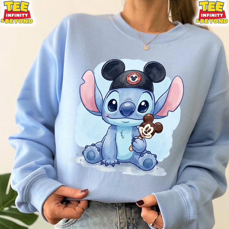 K&ouml;nnte beinhalten: Hellblaues Sweatshirt mit einer Cartoon-Illustration von Stitch, der einen Mickey Mouse-Hut tr&auml;gt und einen Mickey Mouse-Eis am Stiel h&auml;lt. Der Hut tr&auml;gt den Text "Magic Park Adventure". Das Sweatshirt hat den Text "TEE INFINITY & BEYOND".