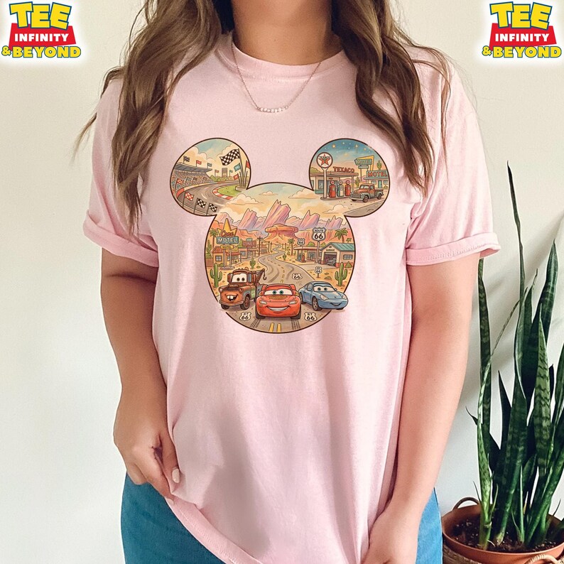 May include: A light pink t-shirt featuring a colorful graphic of the Cars movie characters in a Mickey Mouse head shape. The design includes a race track, desert landscape, and Route 66 elements. The shirt has rolled-up sleeves.