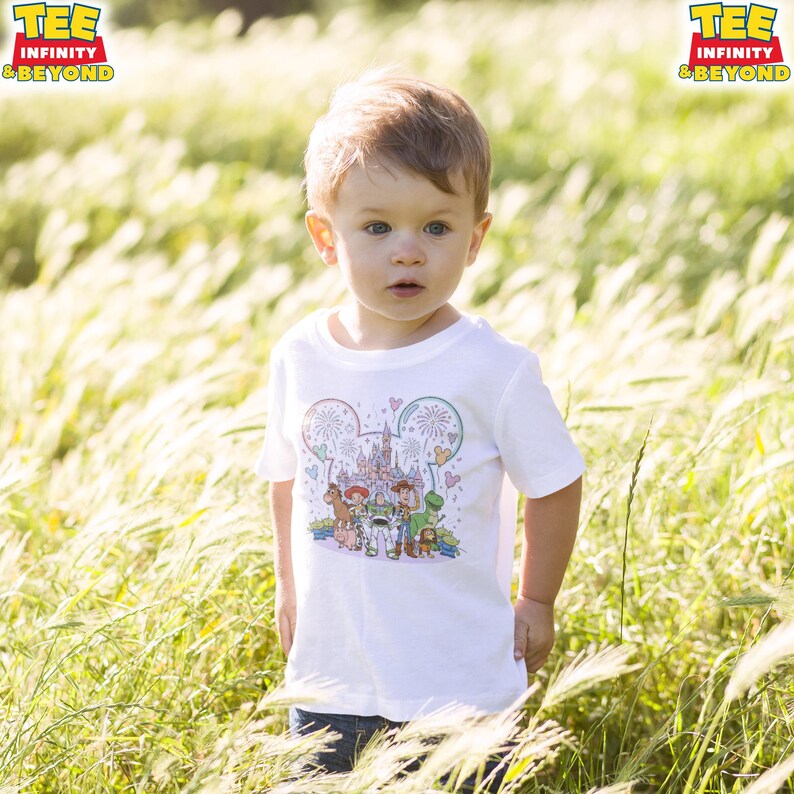 May include: A young child wearing a white t-shirt with a colorful graphic featuring Disney characters and fireworks. The shirt has the text "TEE INFINITY & BEYOND" in a yellow and red logo. The child is standing in a field of tall grass.