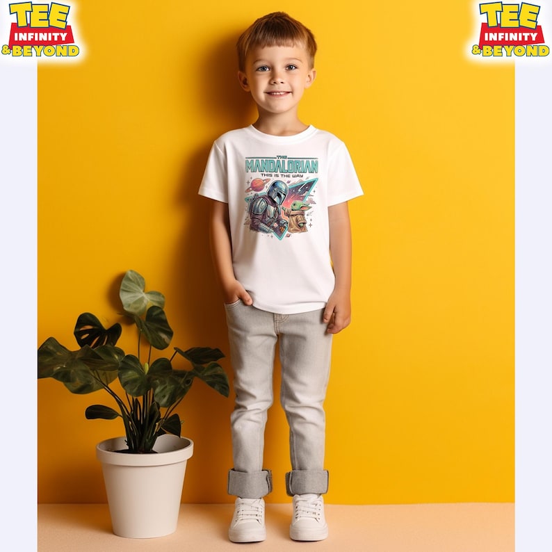 Może przedstawiać: Biały t-shirt z kr&oacute;tkim rękawem z nadrukiem graficznym przedstawiającym Mandalorianina i Grogu oraz napisem "THE MANDALORIAN THIS IS THE WAY" powyżej, na tle o tematyce kosmicznej z kolorowymi planetami i gwiazdami.